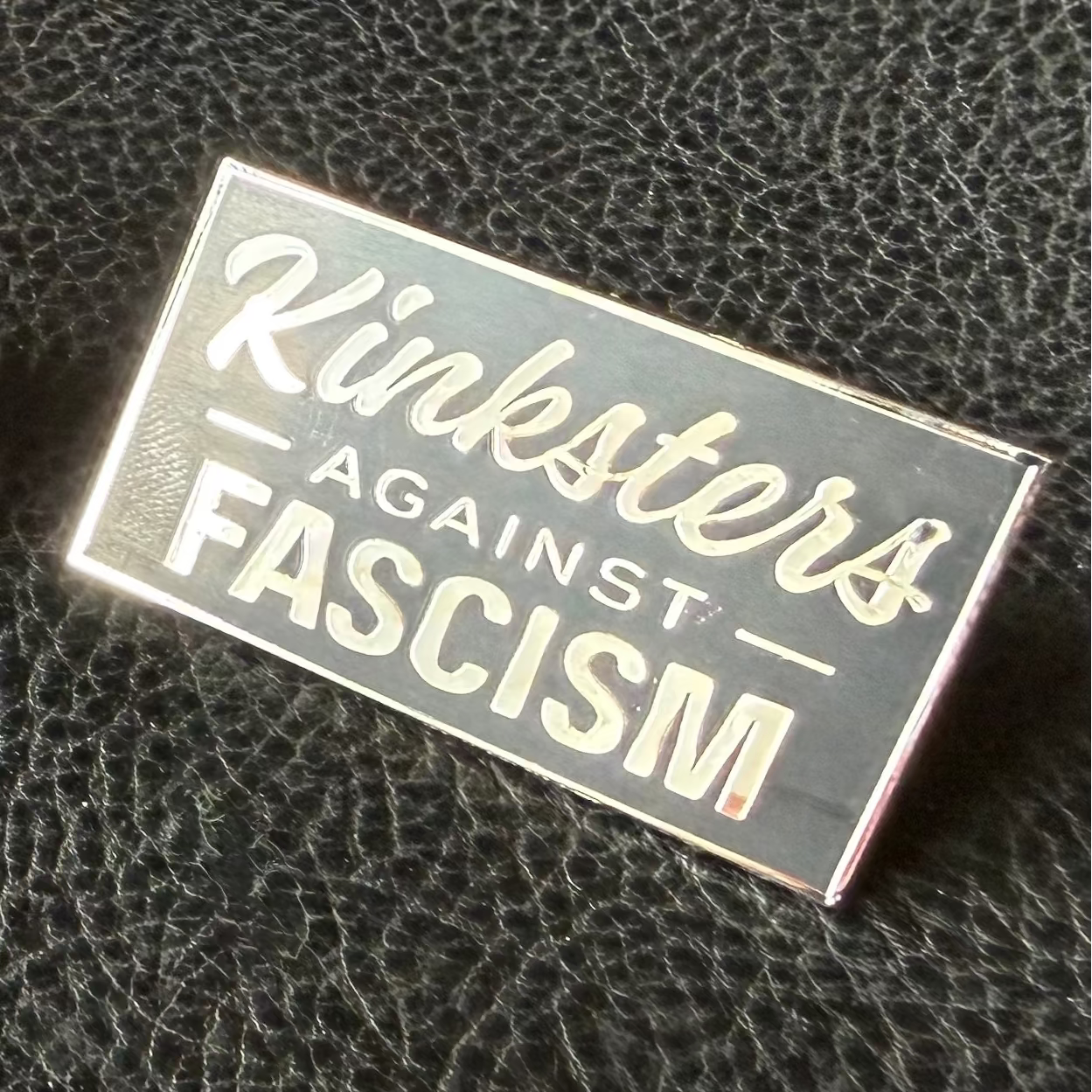 Black enamel pin with 'Kinksters Against Fascism' in silver lettering sitting on a textured black background.